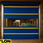 Creative use of roll up doors in livestock and agricultural barns, featuring a blue and yellow Paylon roll-up door with a transparent panel revealing stored agricultural supplies inside a wooden barn