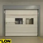 Roll-Up Vinyl Door – Manual by Paylon installed in an industrial warehouse facility, featuring a durable white vinyl curtain with a clear vision panel, set within a concrete block wall.