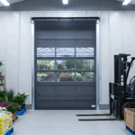 Gray roll-up mesh door open to a greenhouse gardens center with plants, rack, and a forklift nearby