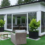 “White pavilion with clear vinyl curtains partially opened, showing a comfortable outdoor lounge space with wicker furniture and greenery,”